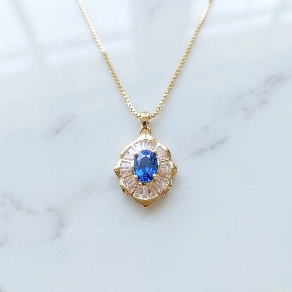 A gold chain necklace with a floral cluster pendant featuring a central blue sapphire surrounded by clear baguette-cut stones in a gold setting, displayed on a white marble surface.