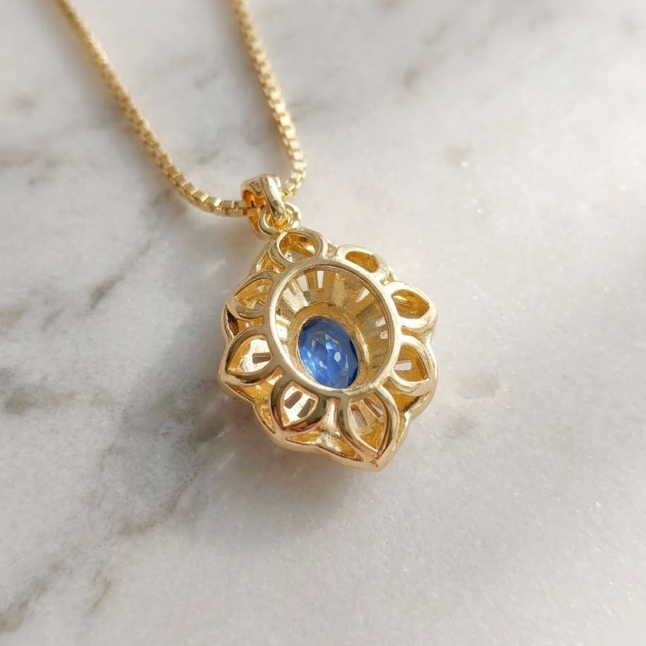 The back of a gold pendant necklace with a open flower design and a central blue oval gemstone, resting on a white marble surface.