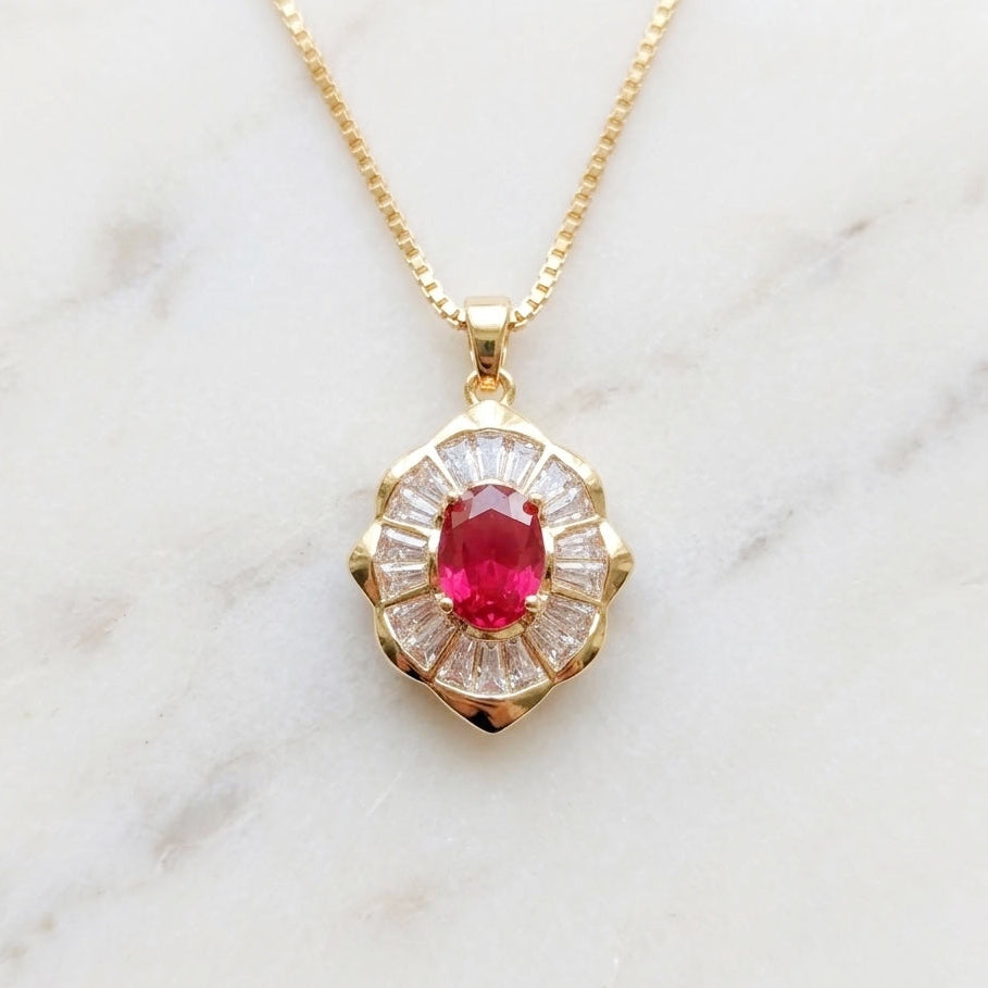 A vintage art deco style gold necklace resting on a marble background. The flower shaped pendant has a red oval cubic zirconia center stone and a halo of clear baguette stones.