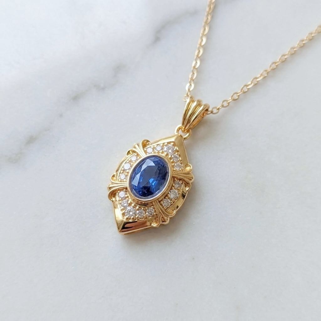 Vintage style gold pendant necklace with a blue oval zirconia stone and small clear accent stones, shown on a white marble background