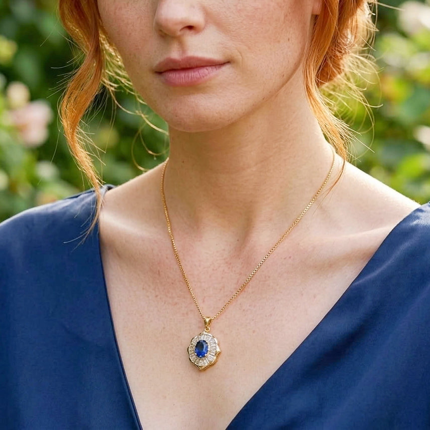 A close-up lifestyle shot of a woman wearing a gold pendant necklace with a blue oval center stone and small clear stones around it, set against a soft green garden background.