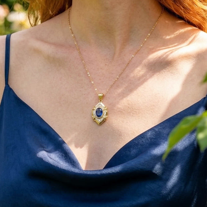 A gold pendant necklace with a blue oval stone and small clear stones, worn by a model in a navy blue satin top.