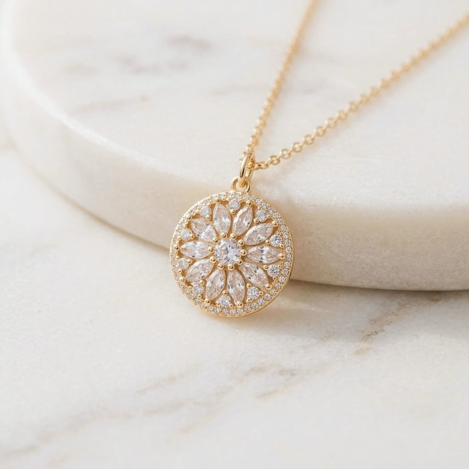 A gold circular pendant necklace with a floral sunflower sunburst design set with clear stones, resting on a white marble surface.