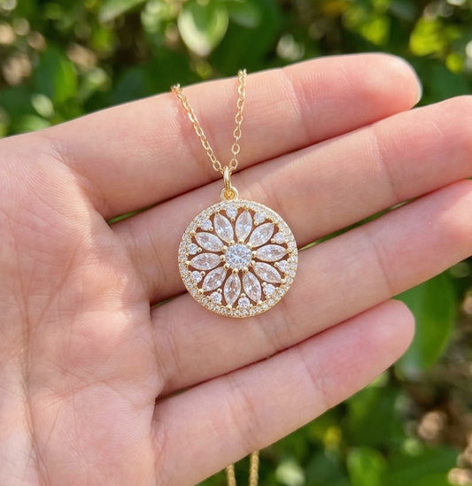 A gold circular mandala pendant necklace with a flower, sunflower or sunburst pattern of clear stones, held in a person's hand against a blurred green outdoor background.