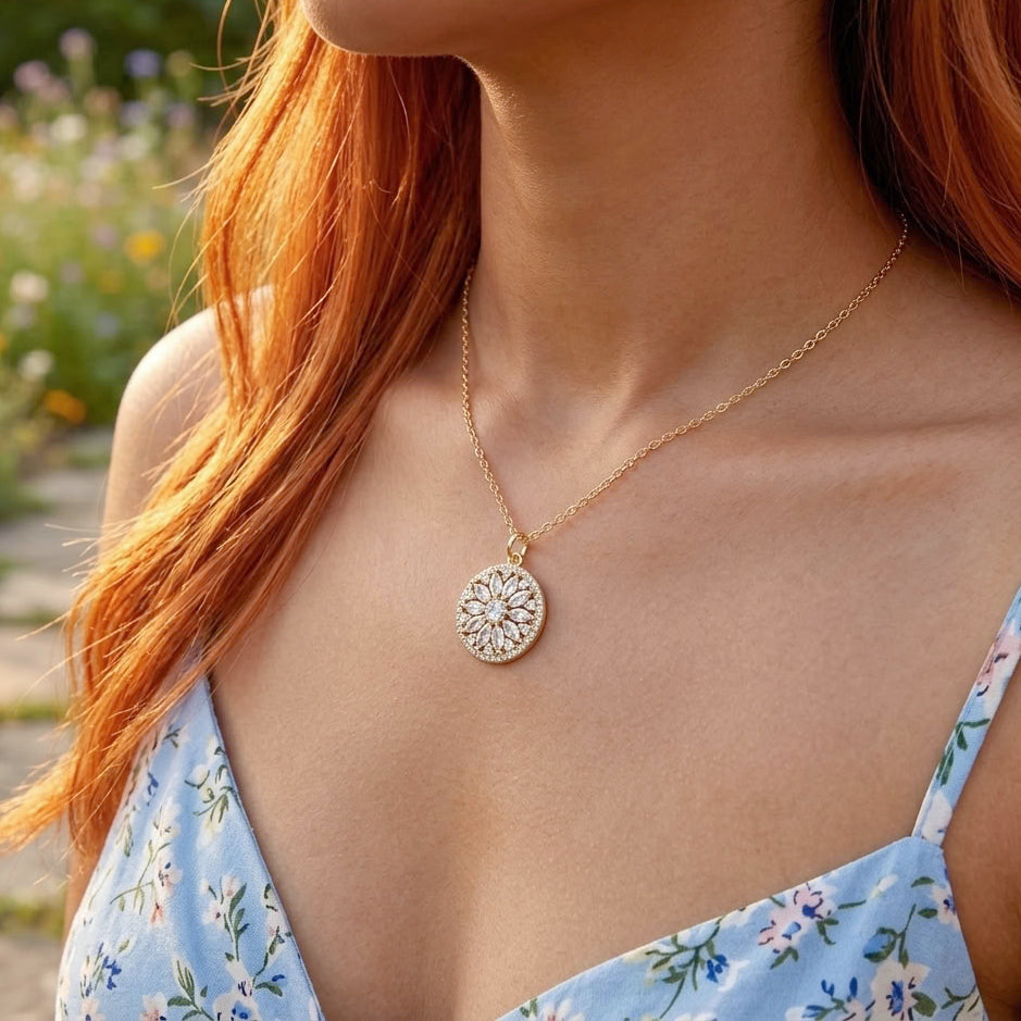A close-up lifestyle shot of a woman wearing a delicate gold chain necklace with a circular, sunburst-patterned pendant. She has vibrant red hair and is wearing a light blue floral top, set against a softly blurred outdoor garden background.