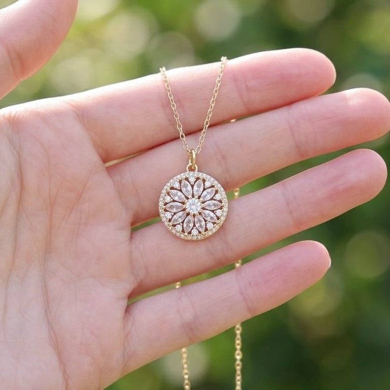 A gold circular pendant necklace with a floral mandala pattern and clear stones, held in a person's hand against a soft green bokeh background.