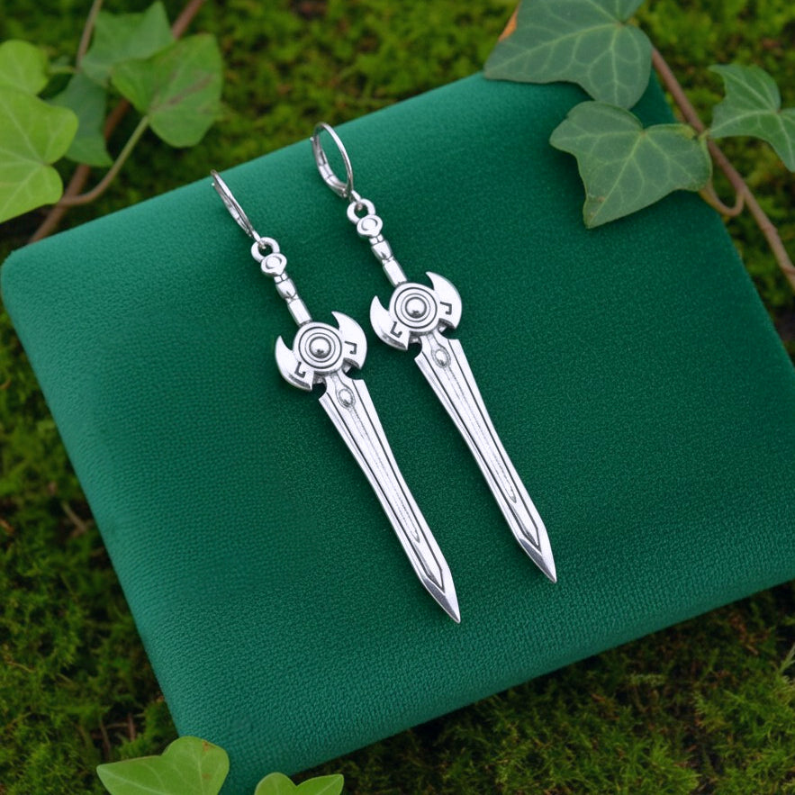 Silver sword earrings on a green textured surface with leaves in the background
