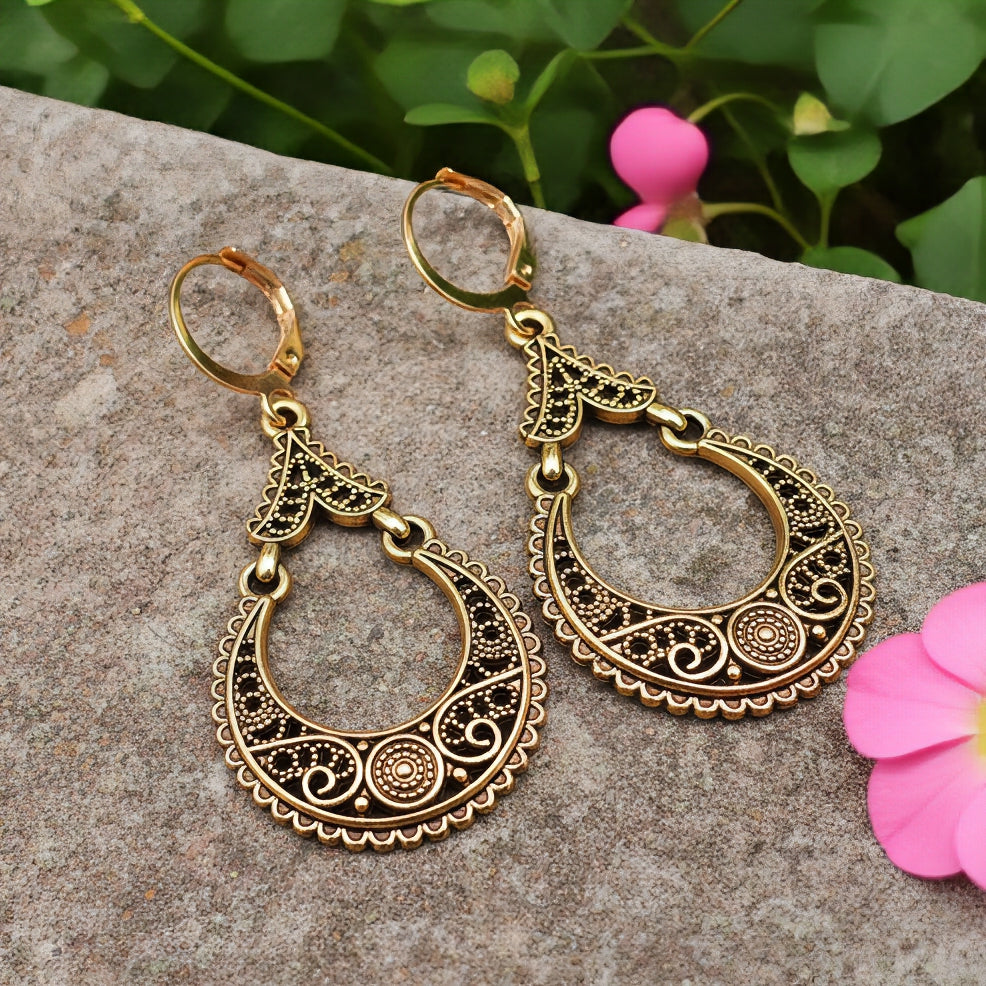Gold hoop earrings with intricate designs on a stone surface with pink flowers and green leaves in the background.