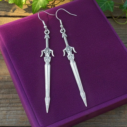 Fantasy style silver sword earrings on a purple velvet background with light coming in
