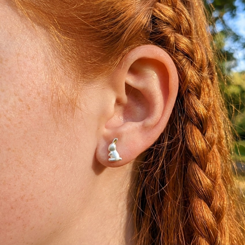 Close-up of an ear wearing a small white bunny-shaped earring with braided red hair.