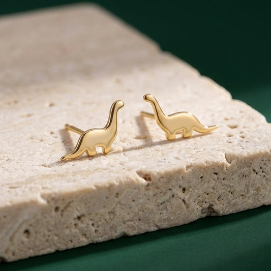 Gold dinosaur-shaped silver stud earrings on a beige stone surface with a green background