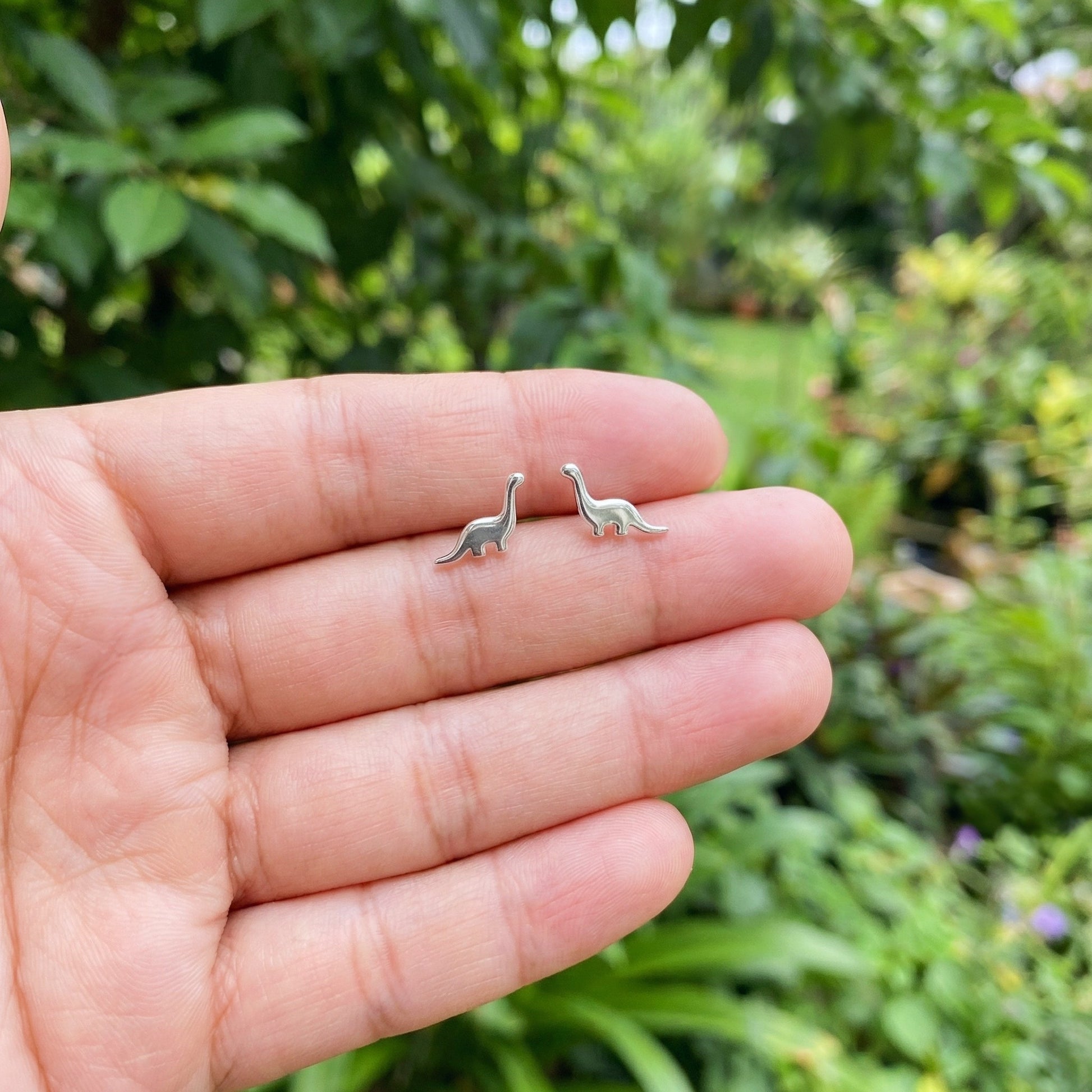 Silver dinosaur earrings held between fingers with a blurred green outdoor background