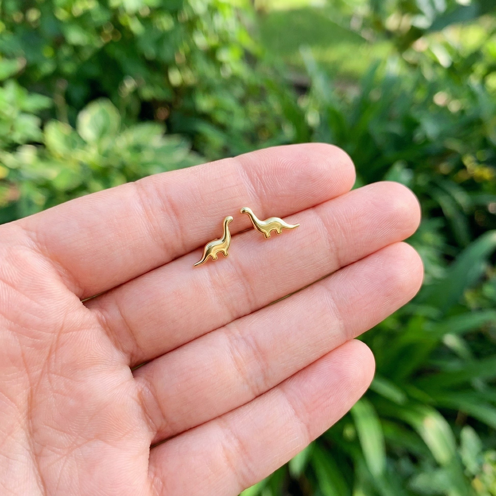 Gold dinosaur-shaped earrings held between fingers with a green outdoor background