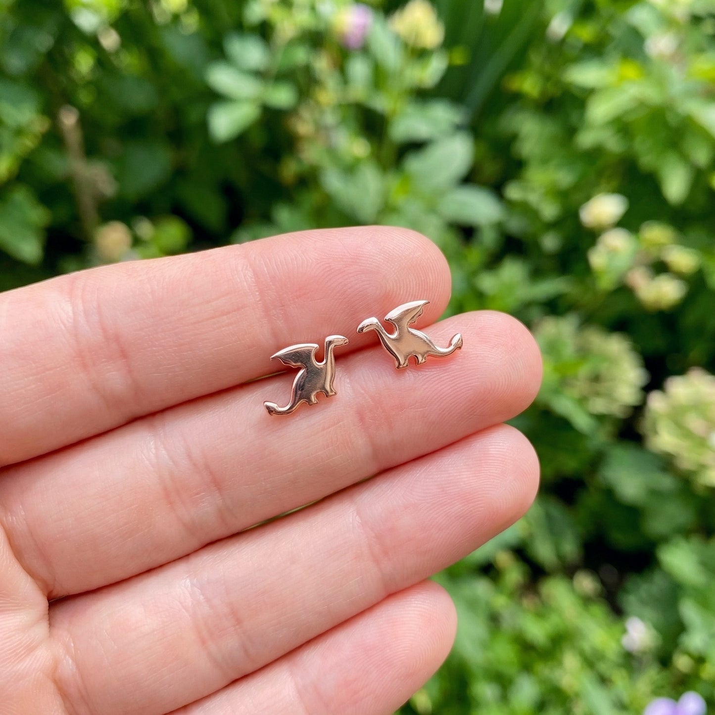 Small rose gold dragon-shaped earrings held between fingers with a blurred green background