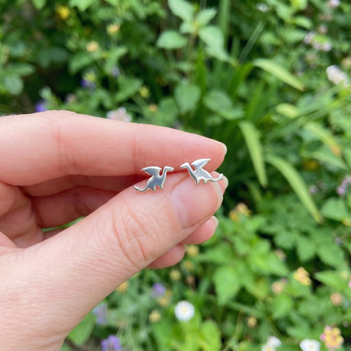 Hand holding two small silver dragon-shaped earrings against a green natural background