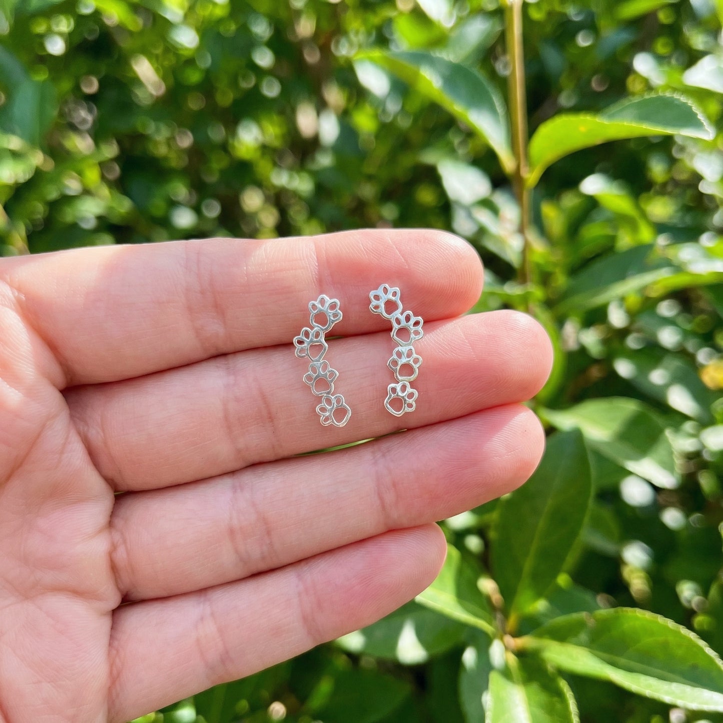 Silver paw earrings held between fingers with a green leafy background