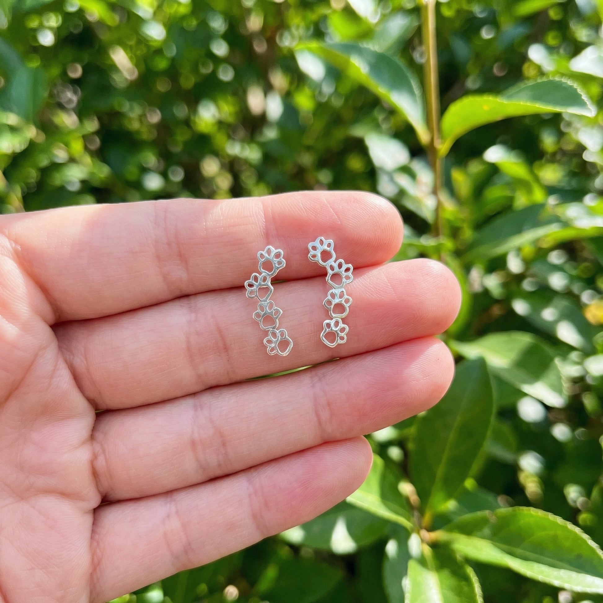 Silver paw earrings held between fingers with a green leafy background