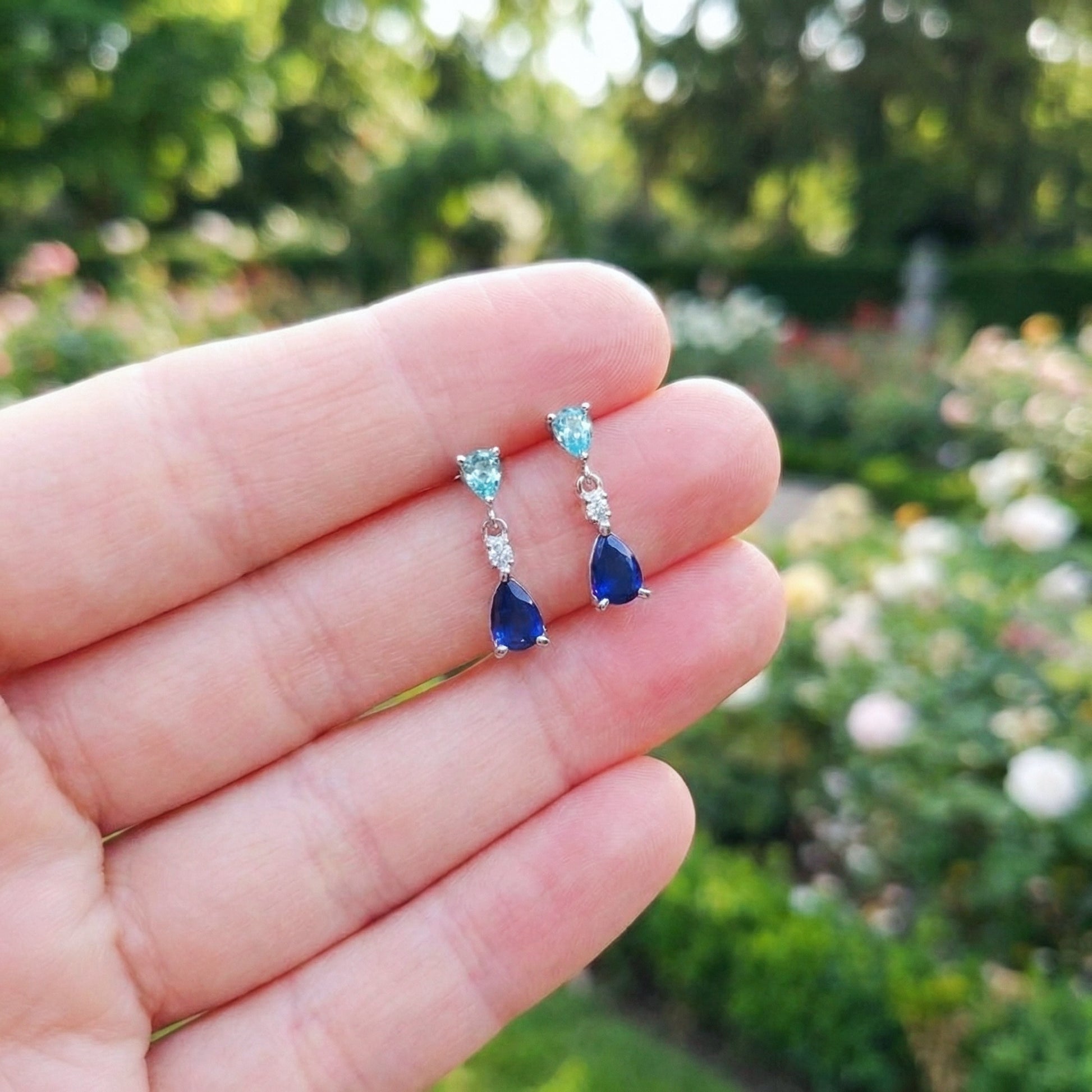 Blue teardrop earrings held in a hand with a blurred garden background
