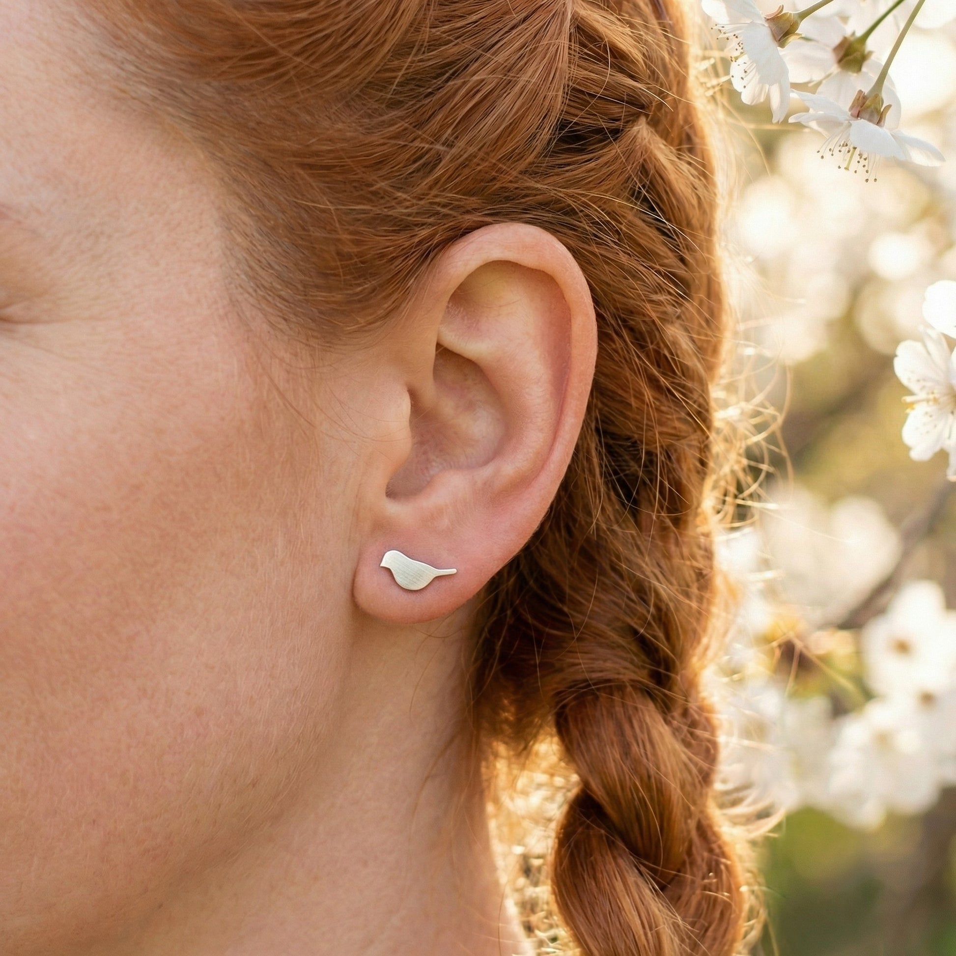 A woman with red hair wearing a silver bird stud earring with cherry blossom behind