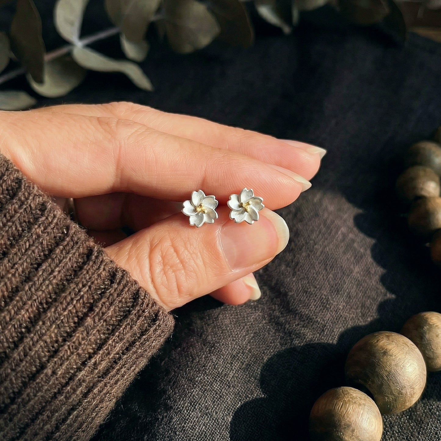 Silver flower earrings held between fingers with a dark fabric background