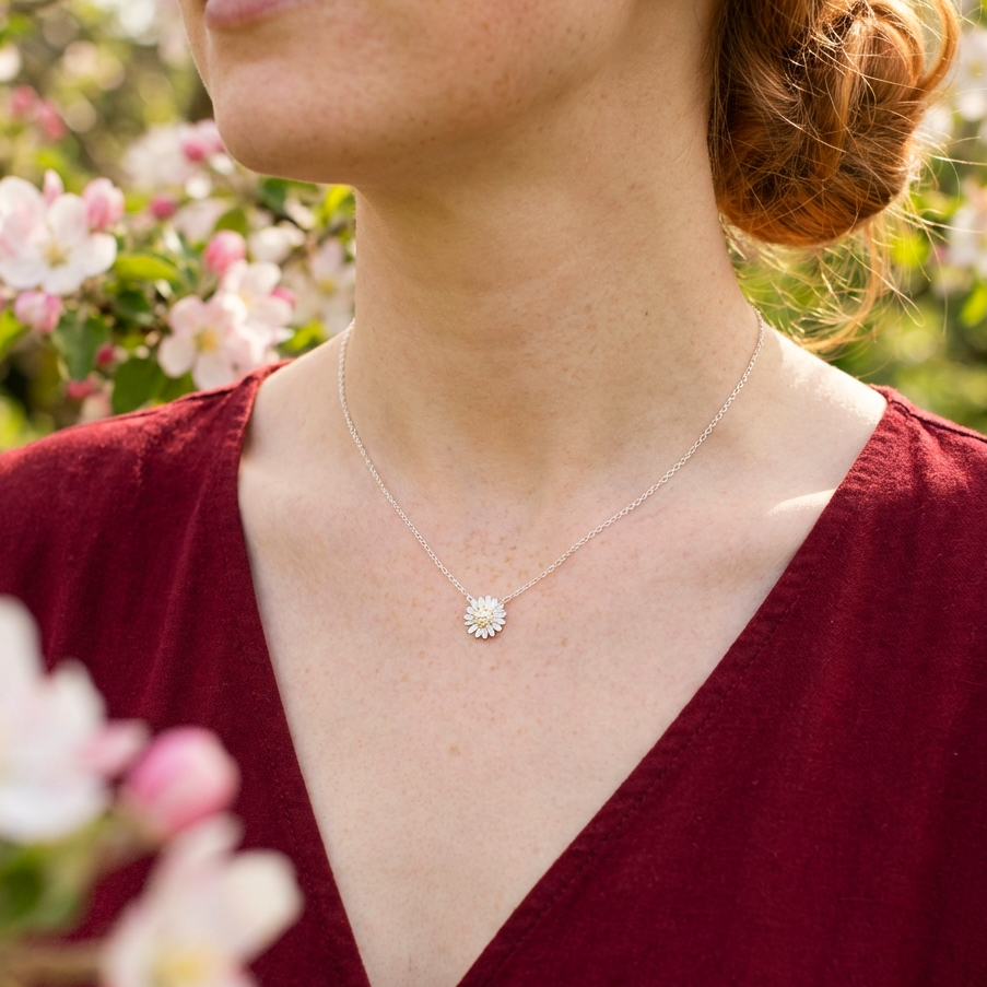 Woman wearing a silver necklace with a daisy pendant, standing in a garden with flowers.
