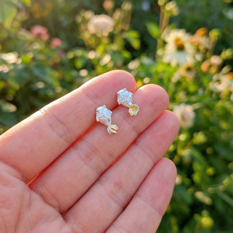A hand holding Silver fish stud earrings with gold tails outside