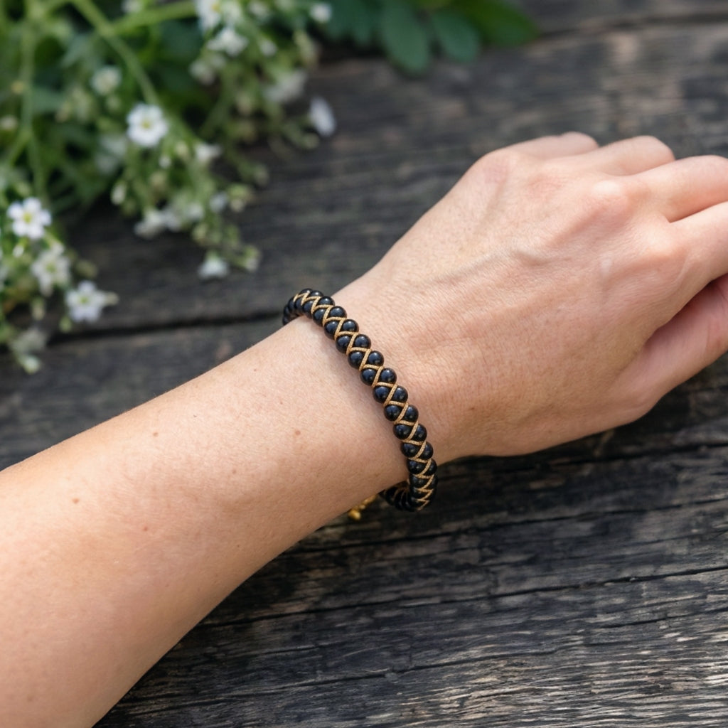 A woman's hand wearing a black obsidian and gold bracelet on a wooden surface with greenery in the background