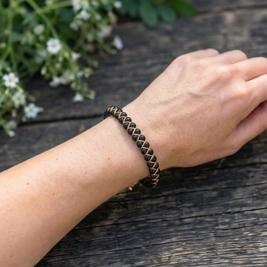 A woman's hand wearing a black obsidian and gold bracelet on a wooden surface with greenery in the background