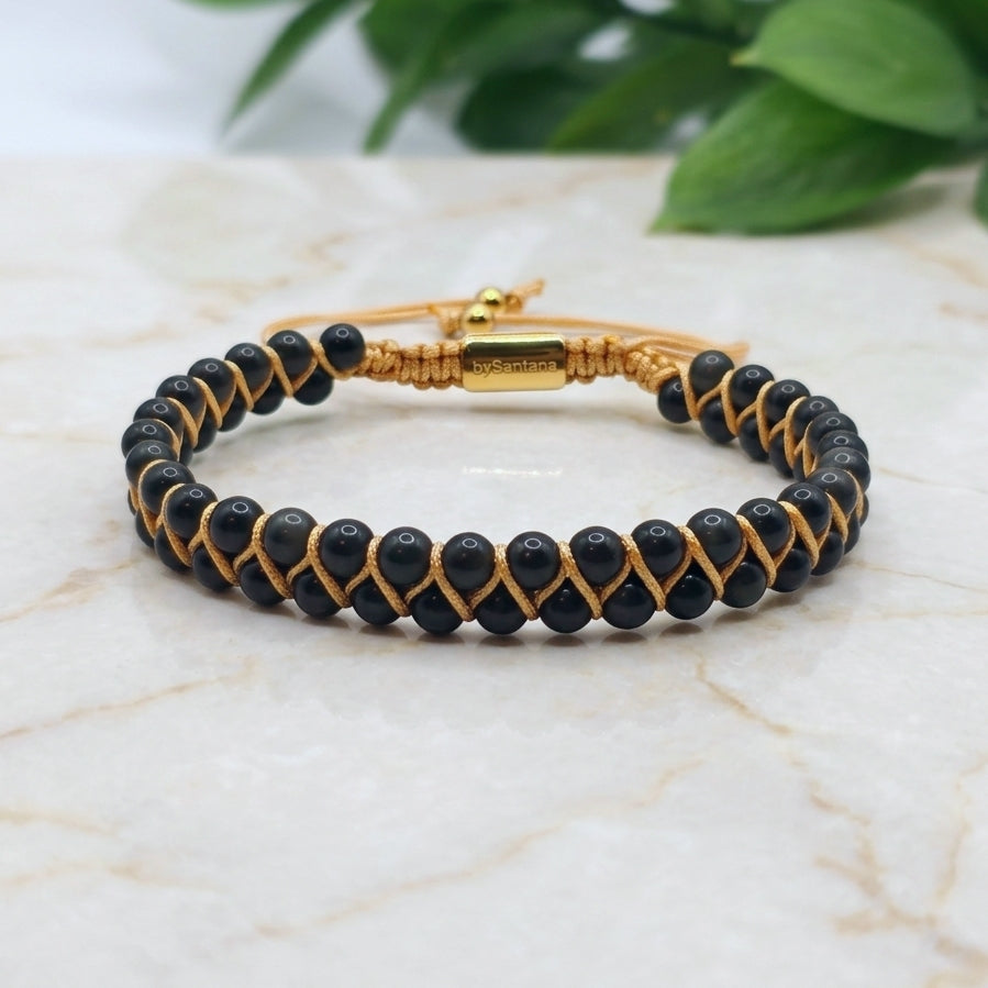 Black obsidian beaded gemstone bracelet with gold accents on a marble surface with green leaves in the background