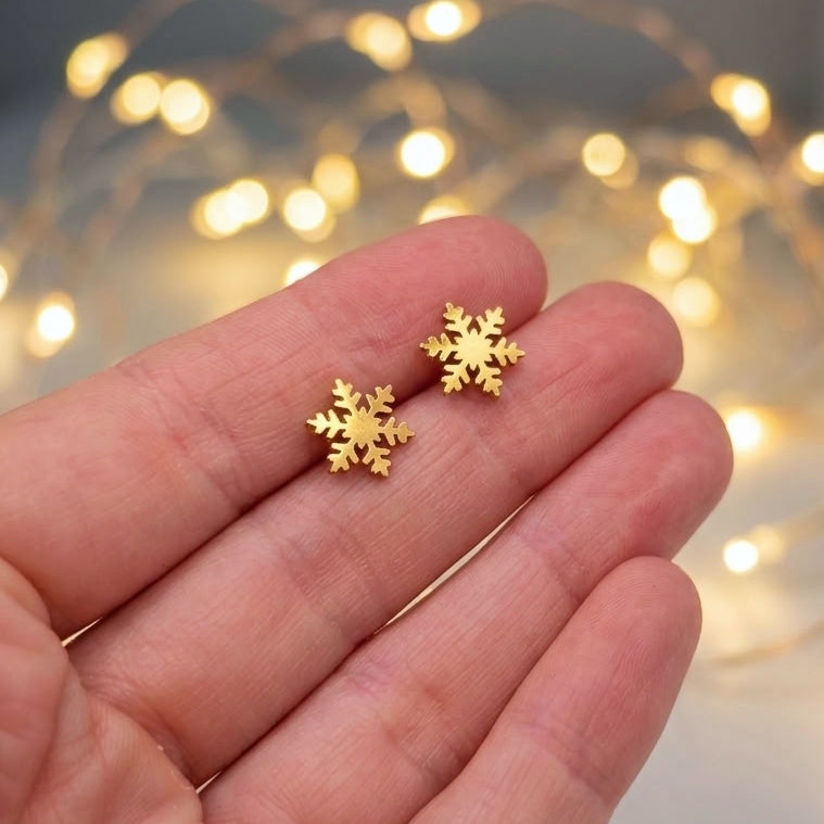 A pair of small gold snowflake stud earrings held on a person's fingers, with warm blurred fairy lights in the background.