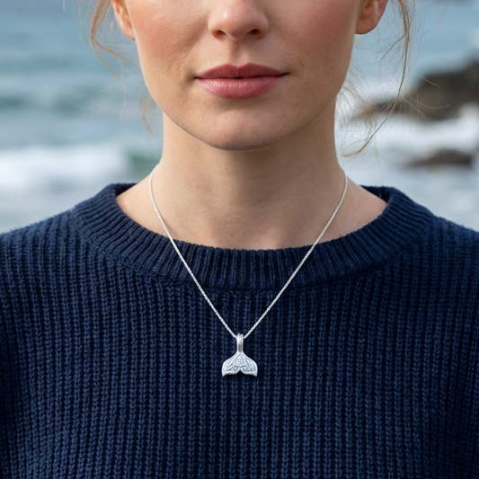 A woman by the sea wearing a silver whale tail necklace on top of a dark blue jumper