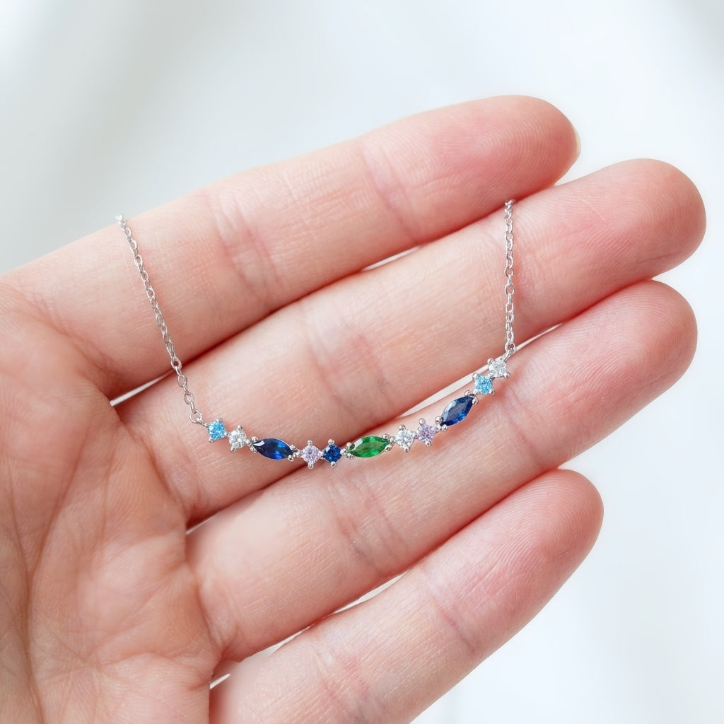 A hand holding a dainty 925 silver bar pendant with blue and green gemstones in marquise and round shapes. 