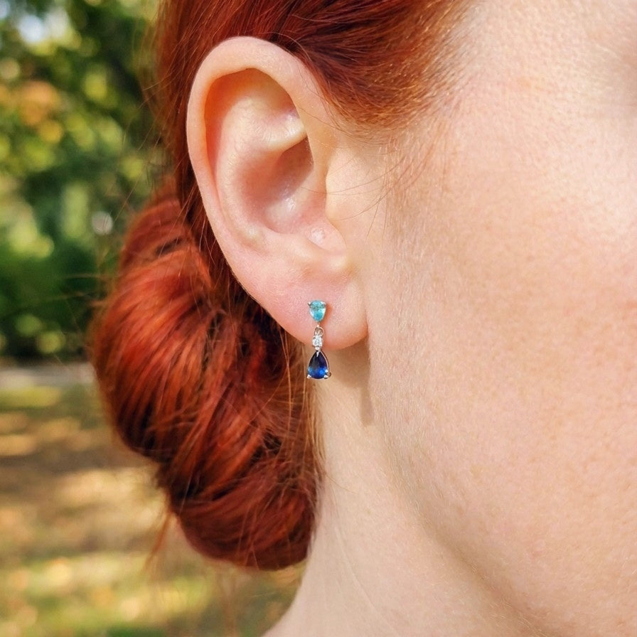 Close-up of an woman's ear wearing a dainty blue gemstone earring with a blurred outdoor background. she has red hair tied back.