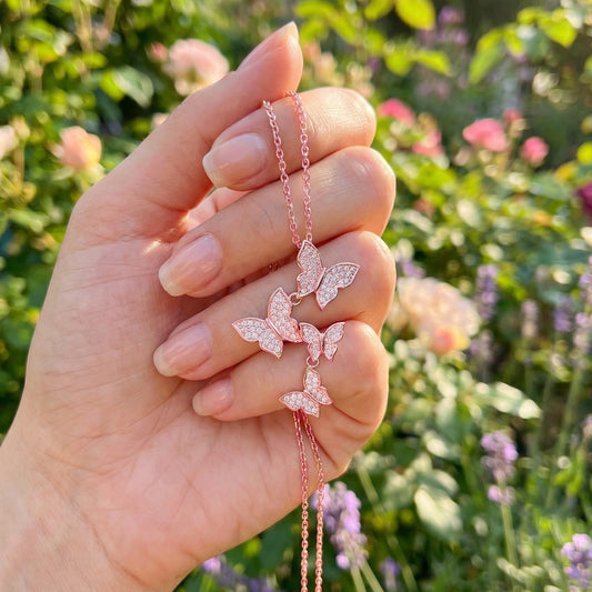Hand holding a delicate pink butterfly necklace against a blurred floral background