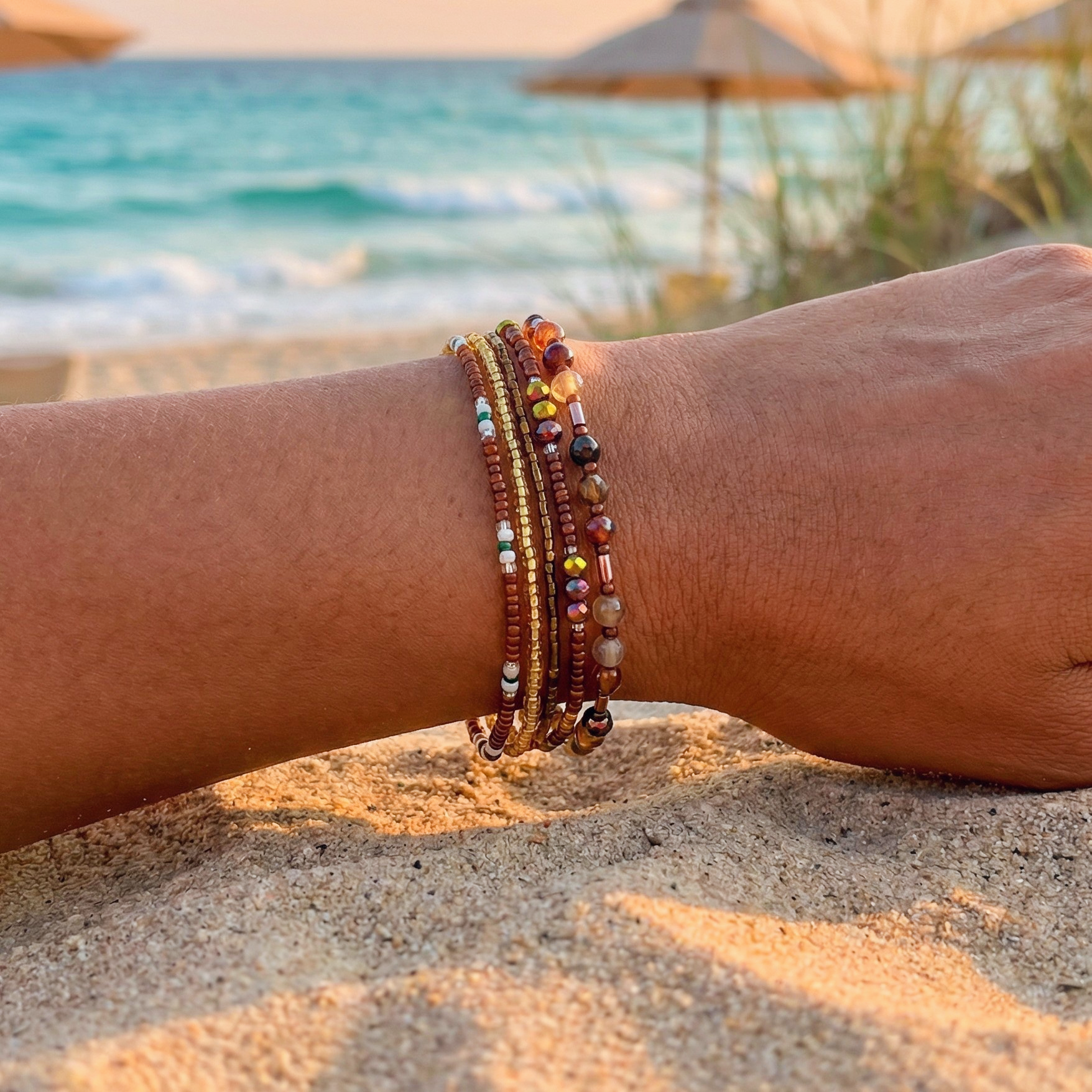 A wrist wearing a multiple bead bracelet with gold and brown beads on a beach