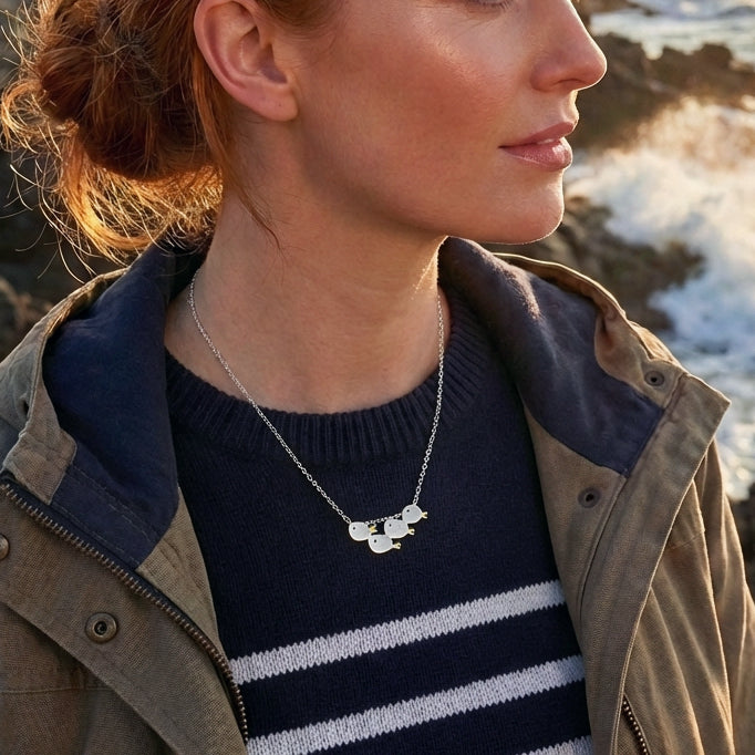 A woman wearing a matte brushed silver necklace with a school of four fishes, standing by the sea, nautical style.