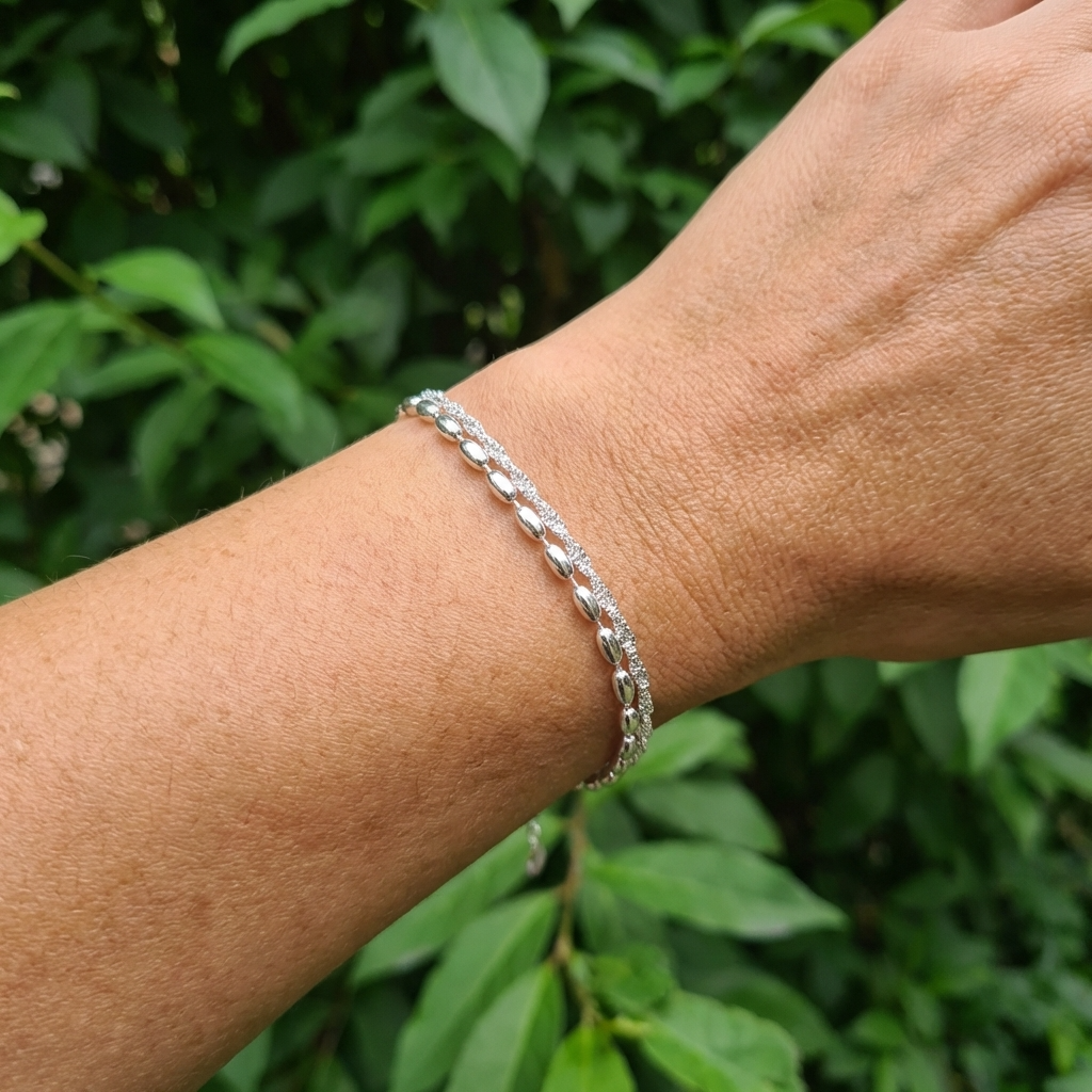 A wrist wearings a double 925 Silver Bracelet featuring one twisted sparkle chain and one with oval beads, outside with greenery behind