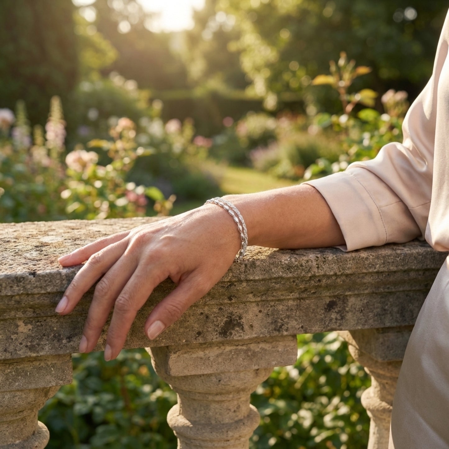 An elegant woman wearing a 925 Silver Bracelet featuring a double chain, one twisted sparkle chain and one with oval beads. She stands by an old stone wall in a fancy garden lit by sunlight.