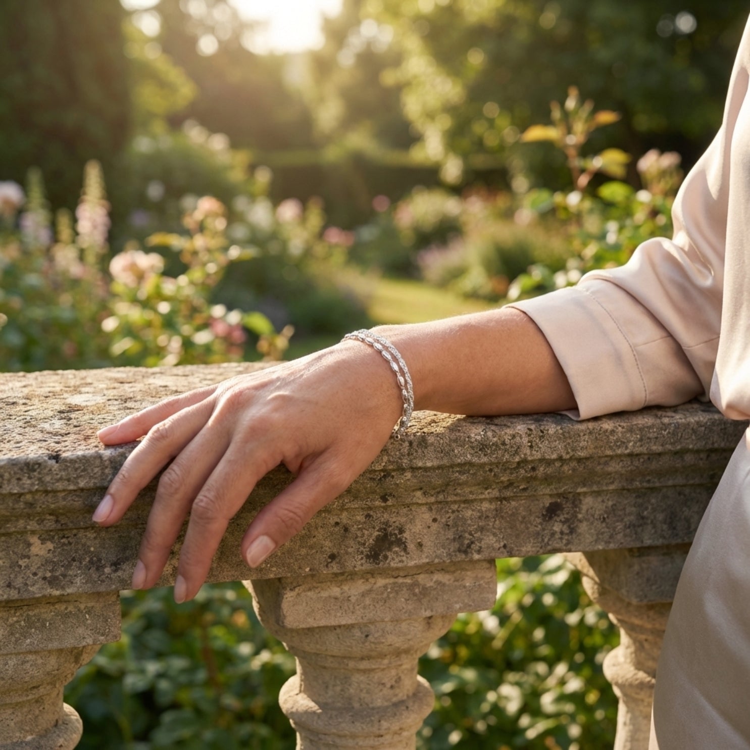 An elegant woman wearing a 925 Silver Bracelet featuring a double chain, one twisted sparkle chain and one with oval beads. She stands by an old stone wall in a fancy garden lit by sunlight.