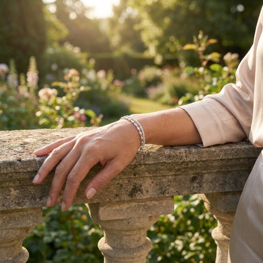 An elegant woman wearing a 925 Silver Bracelet featuring a double chain, one twisted sparkle chain and one with oval beads. She stands by an old stone wall in a fancy garden lit by sunlight.