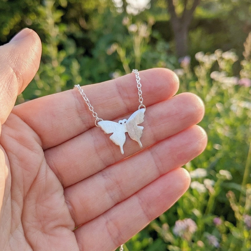 Butterfly-shaped silver pendant on a chain held in a hand with a natural background