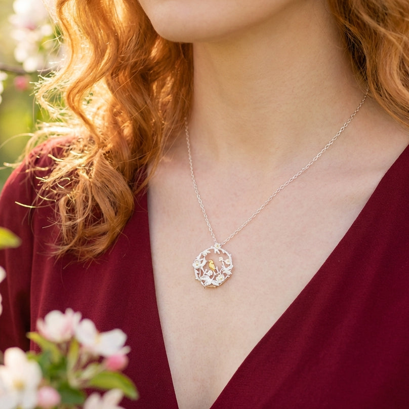 Person wearing a silver necklace with a bird and flower pendant, surrounded by flowers.