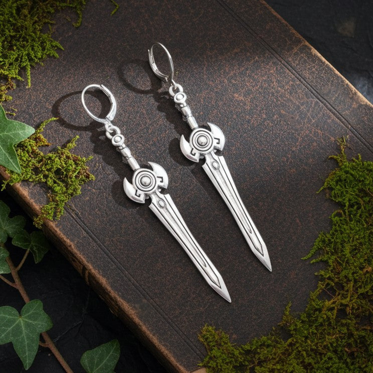 Silver sword earrings with hoops on a dark surface with green leaves.