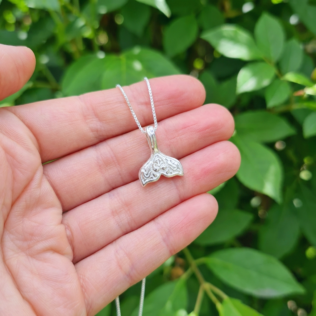 A hand holding a silver whale tail necklace against a green background
