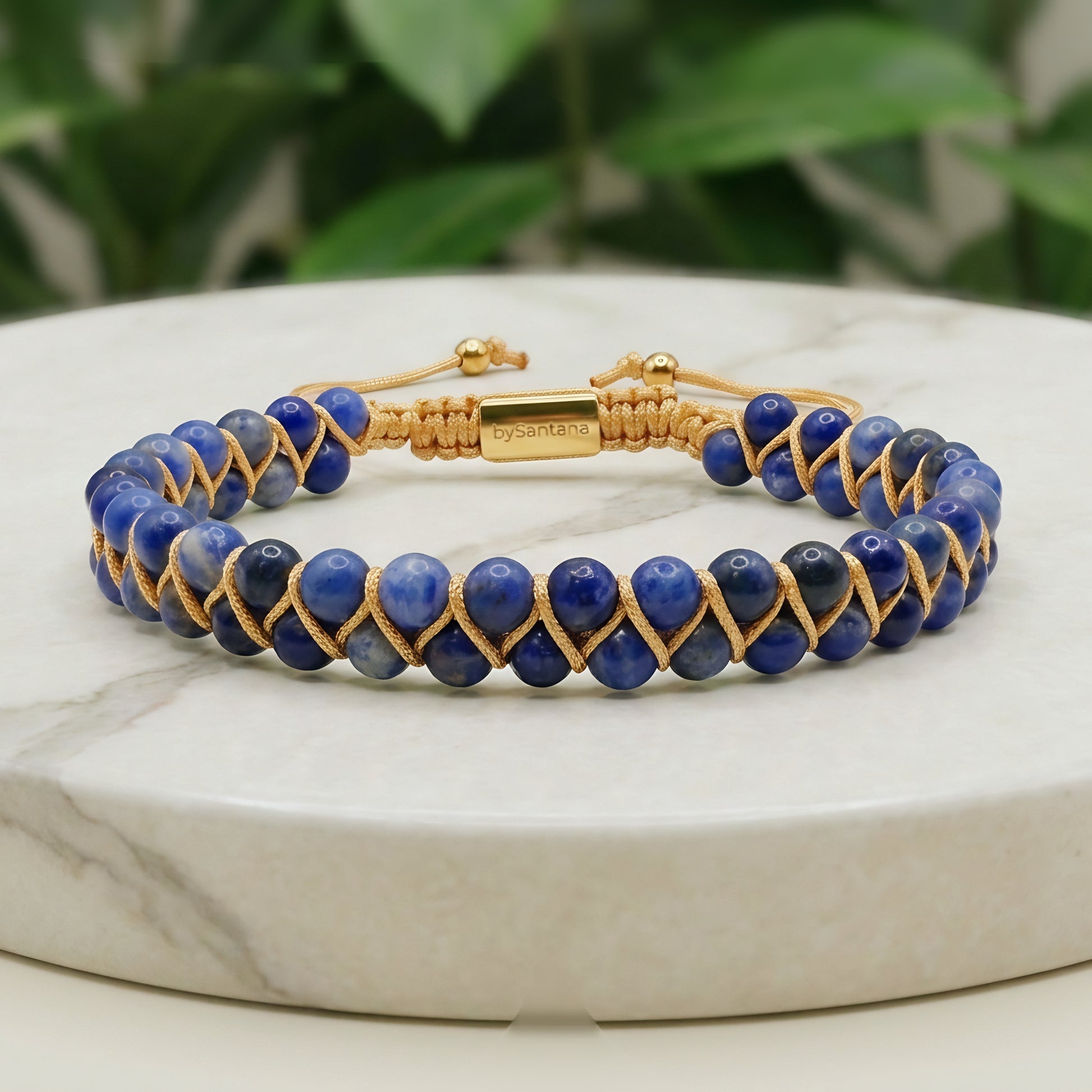 Blue sodalite beaded gemstone bracelet with gold cord on a marble surface with greenery in the background
