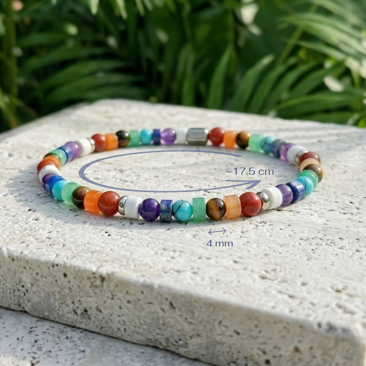 A multicolor beaded bracelet displayed on a light stone surface with green foliage blurred in the background. Blue measurement annotations indicate a circumference of approximately 17.