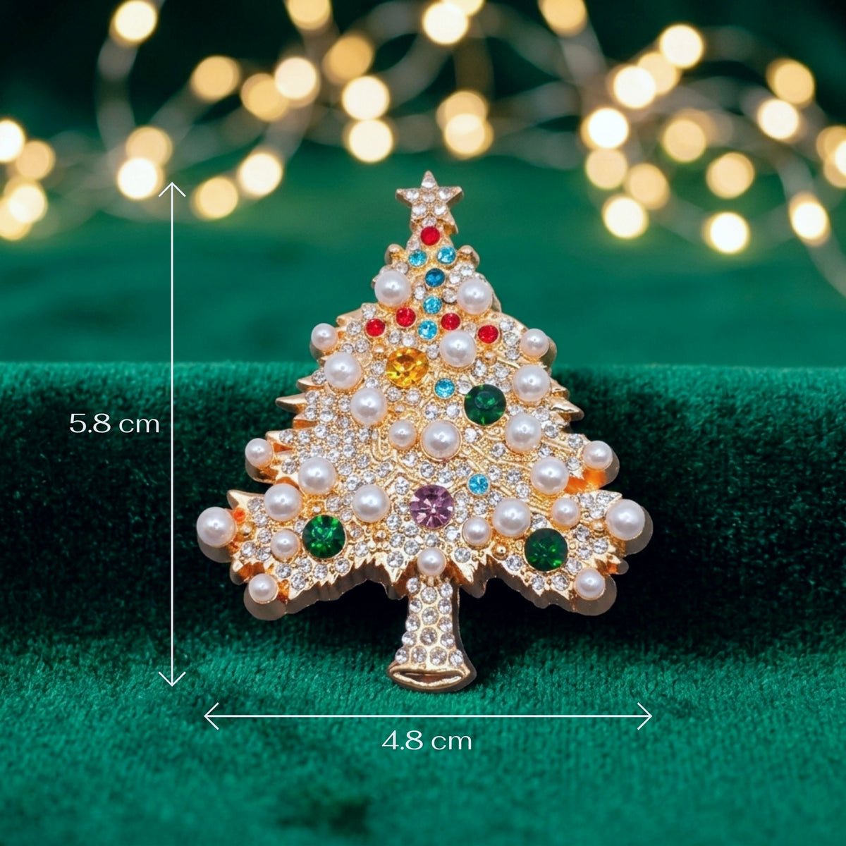 A Christmas tree-shaped brooch with white faux pearls and colorful rhinestones, shown on a green velvet surface with blurred warm string lights in the background. White measurement arrows and text indicate 5.