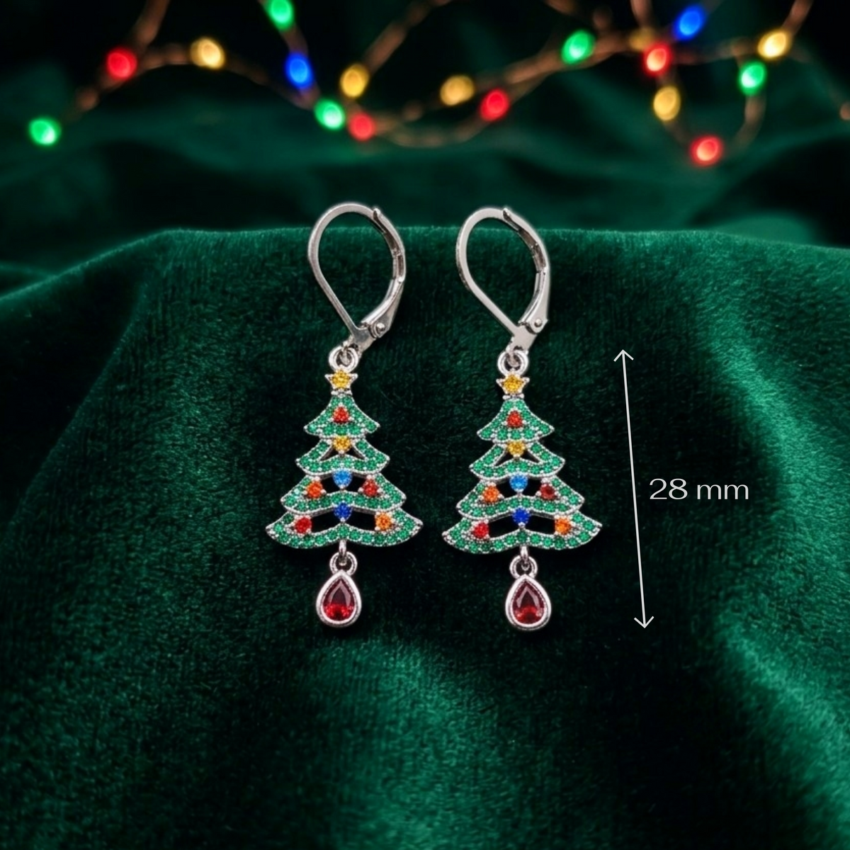 A pair of silver leverback earrings shaped like Christmas trees, decorated with green, blue, red, and yellow cubic zirconia crystals, displayed on a dark green velvet fabric with blurred holiday lights in the background. A vertical dimension marker on the right indicates '28 mm'.