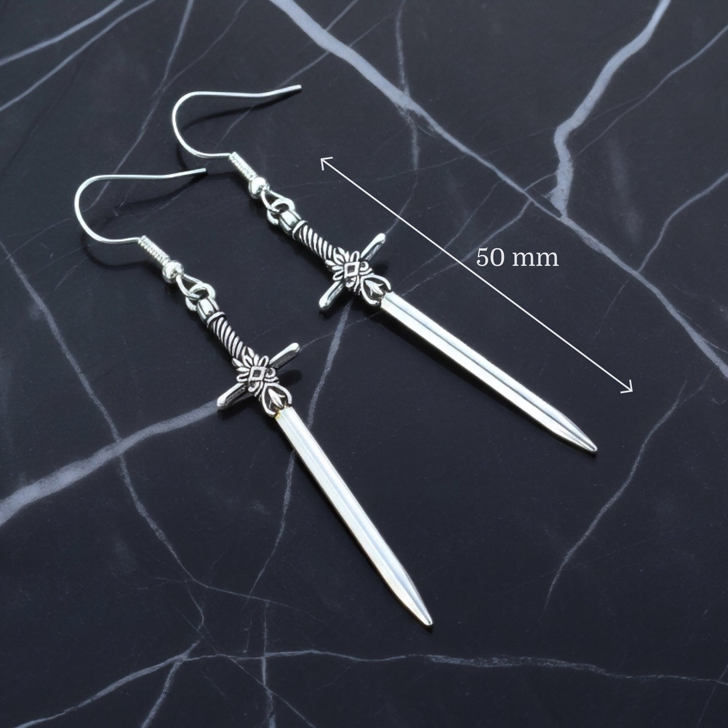Silver sword earrings on a dark marble surface with measurement label.