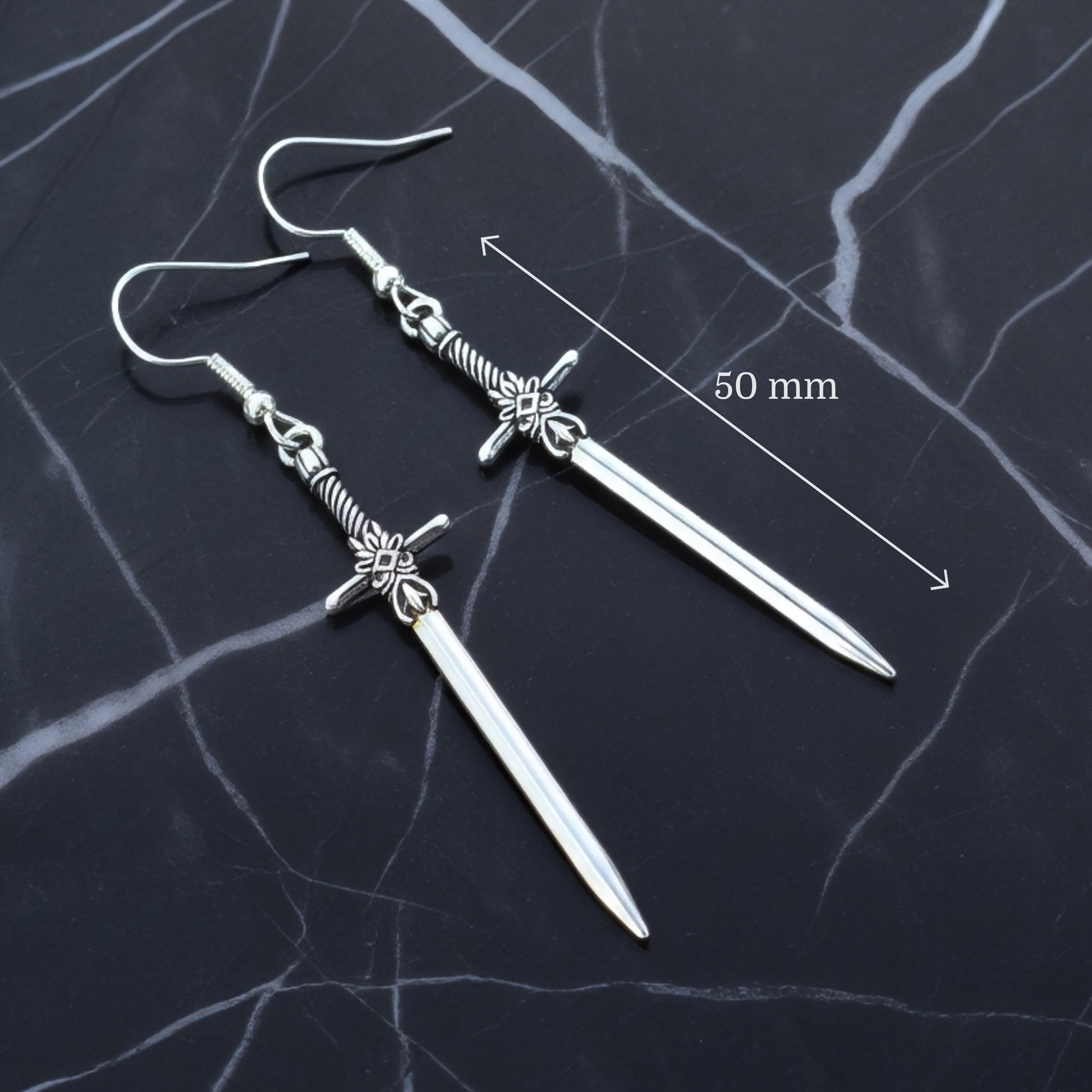 Silver sword earrings on a dark marble surface with measurement label.